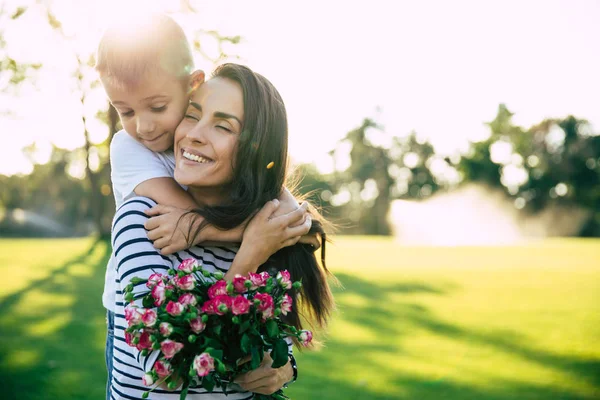 Present on mother\'s day. Little cute son makes surprise with flowers in hands for his happy and beautiful young mother outdoors in park