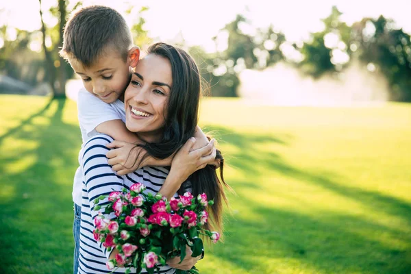 Present on mother\'s day. Little cute son makes surprise with flowers in hands for his happy and beautiful young mother outdoors in park