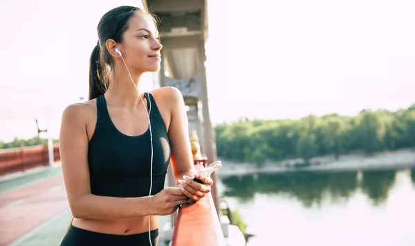 Beautiful and calm young woman in sport clothes is listening music from smart phone and looks on river while stands on the bridge.