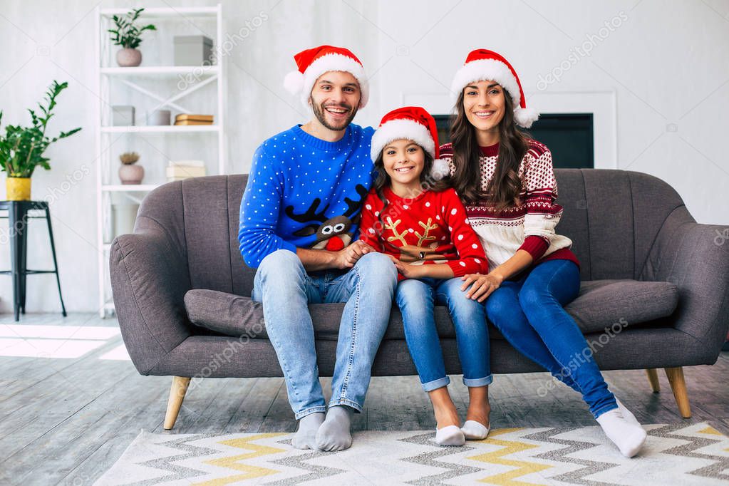 Family of mother, father and daughter in Santa hats and winter sweaters posing for Christmas holiday card of home sofa