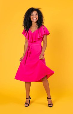 Parlak pembe elbiseli mutlu ırk kadını sarı stüdyo arka planında poz verirken eğleniyor. 