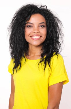Beyaz stüdyo arka planında poz verirken parlak sarı tişörtlü, gülümseyen ve kameraya bakan mutlu, güzel, melez bir kadının portresi. 