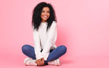 beautiful mixed race woman smiling and looking at camera while sitting cross-legged on pink studio background   clipart