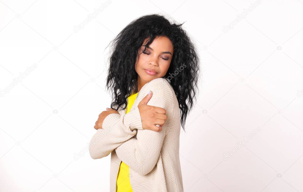 tender beautiful mixed race woman wearing warm clothes hugging herself with closed eyes on white studio background 