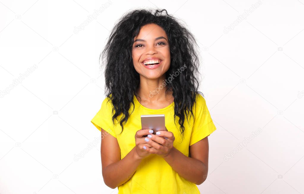 happy mixed race woman in bright yellow t-shirt holding smartphone in hands and looking at camera on white studio background   