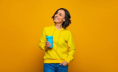 Geleceğe güvenle bakıyorum. Gülümseyen kızın yarım boy fotoğrafı, kendine güvenle yukarı ve yana bakıyor, sağ elinde mavi bir kupa tutuyor, sarı bir kapüşonlu giyiyordu..