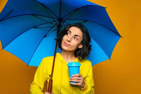 Weather getting better. Half-length photo of pretty girl dressed in a yellow hoodie under an umbrella, holding blue thermal mug in her left hand, glad about weather getting better.