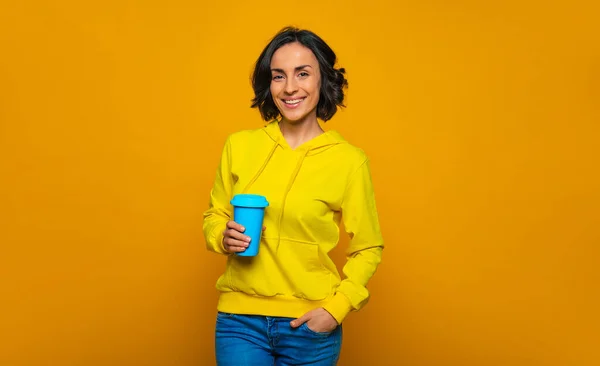 Relaxed day. Half-length photo of smiley relaxed girl dressed in a yellow hoodie, with her left hand in a pocket, looking straight, holding a blue mug in her right hand.