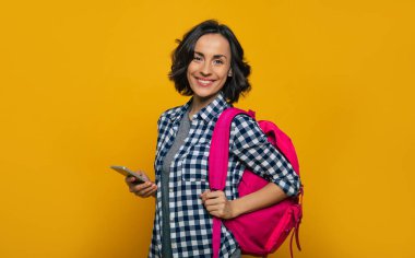 Öğrenci hayatı! Genç ve güzel bir öğrencinin yarım boy fotoğrafı, gündelik giyinmiş, bir omzunda sevimli, pembe bir sırt çantasıyla, geniş bir gülümsemeyle, elinde akıllı telefonunu tutarken..
