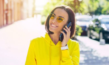 Konuşalım biraz. Sarı kapüşonlu, gözlüklü, elinde akıllı bir bantla modern telefonuyla konuşan güzel bir kızın yakın plan fotoğrafı.