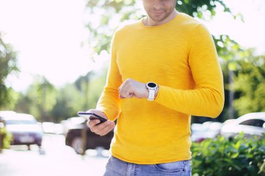 Modern adam. Sarı tişörtlü ve mavi kotlu bir adamın yarım boy fotoğrafı. Saati izliyor, elinde siyah telefonu var..