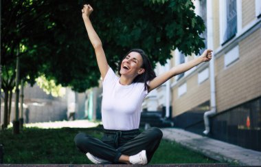Burada olmak çok güzel! Genç bir öğrencinin uzun bir fotoğrafı, dışarıda oturuyor, bacakları çapraz, kolları havada, neşe ve mutluluk hissi yüzünden..