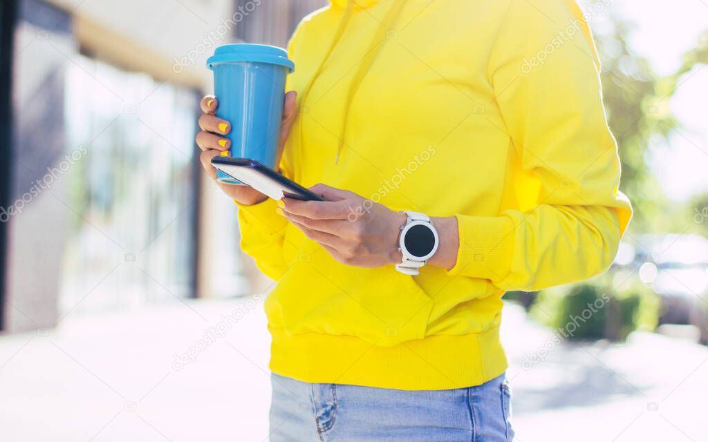 Street style life. A half-length photo of a street style dressed cool woman in a yellow hoodie, with white modern smartwatches on her hand and modern phone in it, holding blue thermal mug.