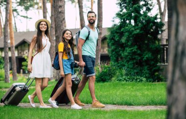 Ormanda bir otel. Ormandaki otellerine doğru yürüyen genç ve mutlu bir ailenin tam boy fotoğrafı. Eşyaları ve geniş tebessümleriyle..