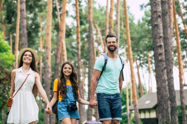 Kırsal kesimde. Mutlu bir ailenin yarım boy fotoğrafı, bavulları ve bavullarıyla ormandaki otellerine giderken, anne ve baba kızlarının elinden tutuyorlar..
