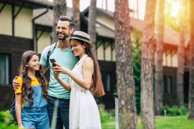 Keyifli yolculuklar. Neşeli, güler yüzlü, gezgin bir ailenin otelin yakınında oturduğu bir fotoğraf, baba, kız ve anneye sarılmış, anne telefona bakıyor..