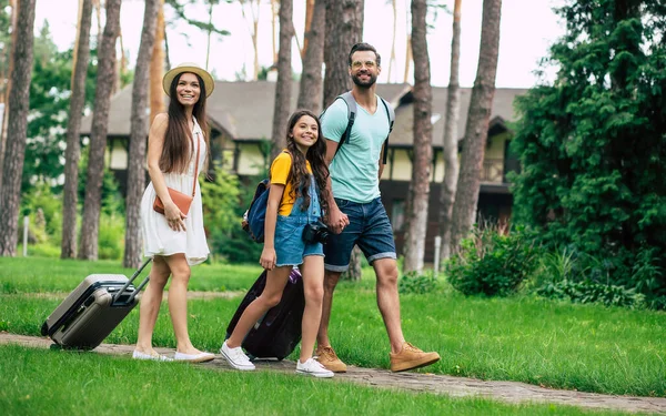 Going to a hotel in the forest. A full-length photo of a happy family, going to their hotel in the forest, with their belongings and wide smiles, while father and daughter are holding hands.