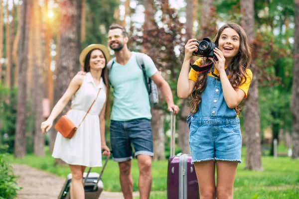 Bunu sen de görüyor musun? Parkta bavulları olan mutlu bir ailenin tam boy fotoğrafı. Tulum giymiş heyecanlı bir kız, fotoğraf çekmeye hazır..