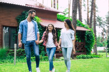 Hoş bir kızı olan genç, mutlu, modern ebeveynler banliyö evinin arka planında el ele tutuşuyorlar..