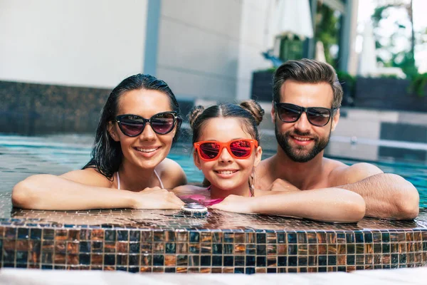 漂亮的微笑的男人和可爱的兴奋的女人和他们可爱的小女儿在宾馆休息的时候 正在夏天的游泳池里玩乐 — 图库照片