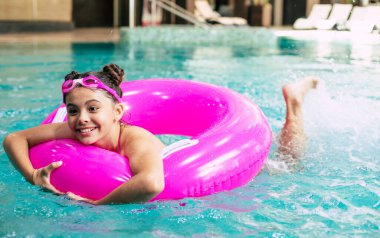 Gözlüklü mutlu küçük kız sıcak bir yaz gününde yüzme havuzunda pembe şişirilebilir bir yüzükle oynuyor. Çocuklar yüzmeyi öğrenir. Çocuk oyuncakları. Aile plaj tatili..
