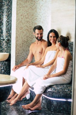 Banyo havlularıyla gülümseyen güzel bir aile hamamda ya da saunada dinlenirken kameraya bakıyor. Tatil ve Spa prosedürü