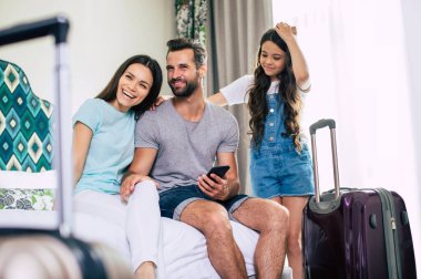 Büyük mutlu ve heyecanlı turist ailesi otel odasındaki yatakta otururken akıllı telefon kullanıyor. İnternette aranıyor