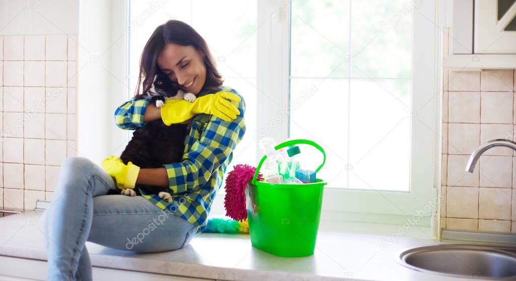 Happy beautiful young woman in yellow gloves is cleaning the kitchen with special equipment and plays with cute cat