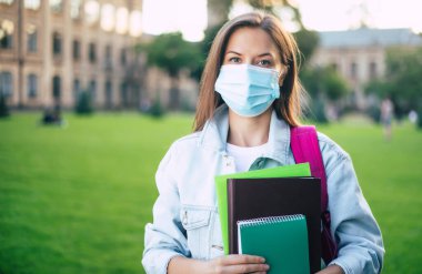 Elinde kitap ve belgelerle tıp maskesini koruyan kendine güvenen genç bayan kameraya bakıyor ve üniversite kampüsünün arka planında duruyor.