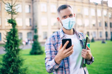 Tıp maskeli genç ve kendine güvenen bir öğrenci kameraya bakıyor ve üniversitenin arka planında duruyor.