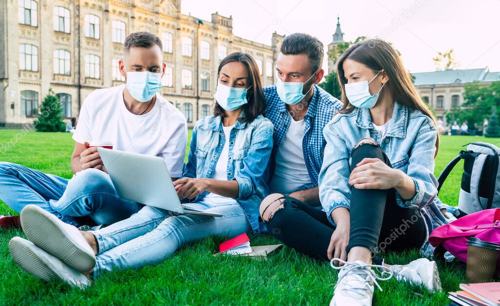 Group of young students in medical masks with laptop and books are studying together in university. Friends outdoors sitting on the grass.
