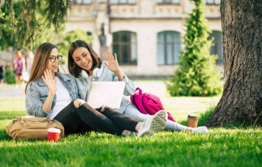Spor kıyafetleri içinde iki mutlu genç kız öğrenci üniversite parkında dizüstü bilgisayar ve akıllı telefonla dinleniyor ve kahve içiyorlar..