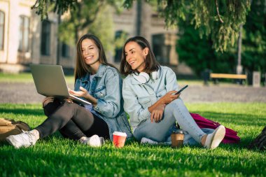 Spor kıyafetleri içinde iki mutlu genç kız öğrenci üniversite parkında dizüstü bilgisayar ve akıllı telefonla dinleniyor ve kahve içiyorlar..