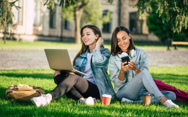 Spor kıyafetleri içinde iki mutlu genç kız öğrenci üniversite parkında dizüstü bilgisayar ve akıllı telefonla dinleniyor ve kahve içiyorlar..
