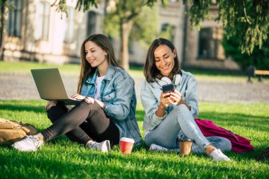 Spor kıyafetleri içinde iki mutlu genç kız öğrenci üniversite parkında dizüstü bilgisayar ve akıllı telefonla dinleniyor ve kahve içiyorlar..
