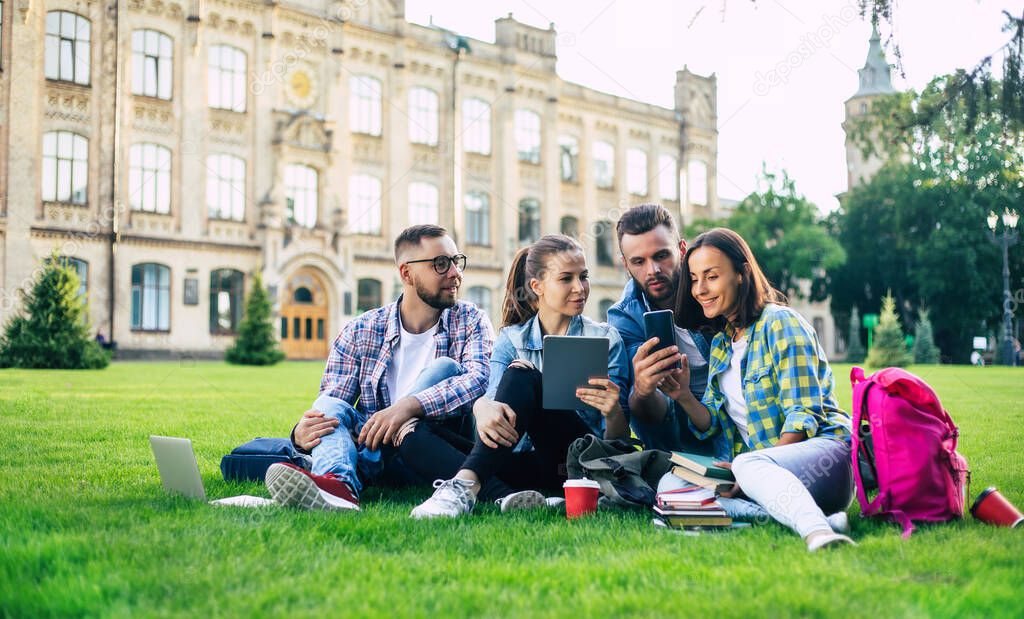 Happy beautiful students team with backpacks and other stuff are talking, having fun and relaxing on the grass on college background outdoors