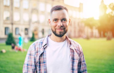 Gündelik giysiler içinde genç ve yakışıklı sakallı bir öğrenci üniversite binasının arka planında poz veriyor ve kameraya bakıyor.