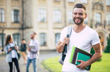 Sırt çantası ve kitapları olan sakallı, genç ve yakışıklı bir öğrenci üniversite binalarında yürüyor ve poz veriyor.