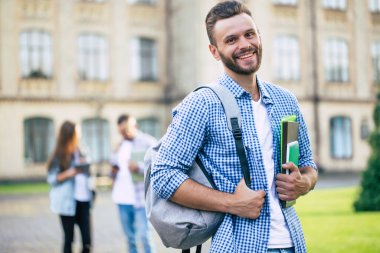 Sırt çantası ve kitapları olan sakallı, genç ve yakışıklı bir öğrenci üniversite binalarında yürüyor ve poz veriyor.