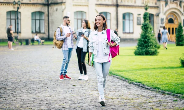 若いです美しいですブルネット学生女の子でデニム服にバックパックと本で手に彼女の学生の友人のグループに立っている背景 — ストック写真