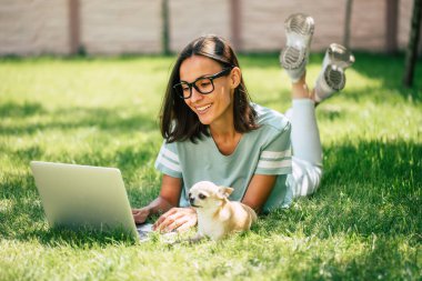 Güzel, mutlu, küçük köpeğiyle serbest çalışan genç bir kadın dışarıda bir dizüstü bilgisayar üzerinde çalışıyor..