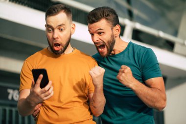 Stadyum arka planında elinde akıllı bir telefonla iddiayı kazandıktan sonra coşkulu ve heyecanlı iki hayran arkadaş.