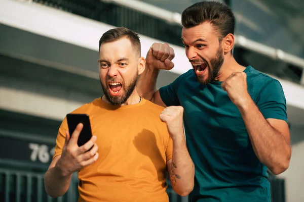 Stadyum arka planında elinde akıllı bir telefonla iddiayı kazandıktan sonra coşkulu ve heyecanlı iki hayran arkadaş.