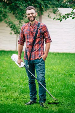 Evin arka bahçesinde çim biçme makinesi olan kareli gömlekli yakışıklı bir bahçıvan.