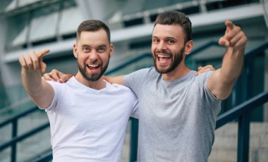 Kamerayı gösteren mutlu adamlar. Renkli tişörtler giymiş iki sakallı yakışıklı arkadaş şehrin arka planında birlikte poz veriyorlar.. 