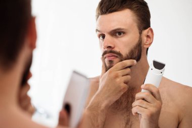 Handsome young man shaving his beard in the bathroom. Portrait of a stylish naked bearded man examining his face in-home mirror.  clipart