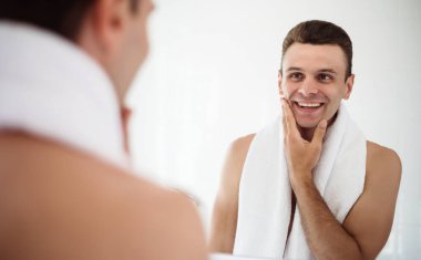Handsome young man shaving his beard in the bathroom. Portrait of a stylish naked bearded man examining his face in-home mirror.  clipart