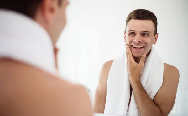 Handsome young man shaving his beard in the bathroom. Portrait of a stylish naked bearded man examining his face in-home mirror.