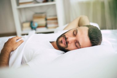 Yakışıklı genç sakallı adam rahat bir yatak odasında beyaz büyük bir yatakta uyuyor.