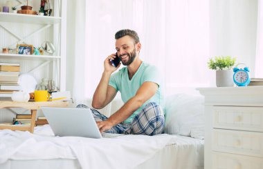 Gündelik giysiler içindeki yakışıklı genç adam yatakta otururken evde çalışıyor. Dizüstü bilgisayarı ve akıllı telefonu olan kendine güvenen bir adam yatak odasında kahve içer..